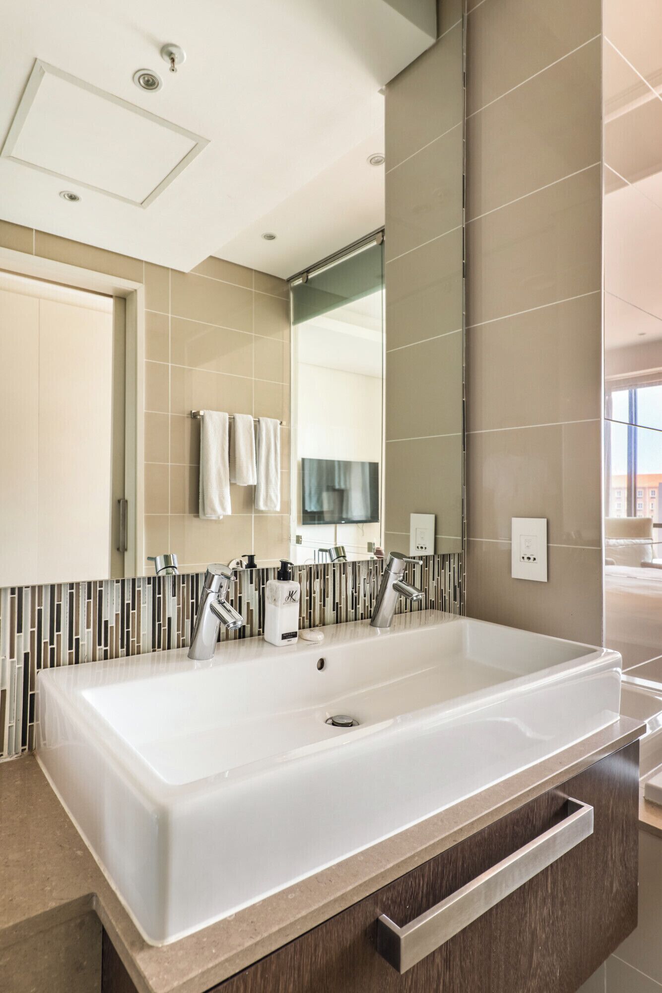 A modern bathroom with a large white sink, two faucets, a mirror, and a tiled backsplash.