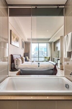 Ett modernt hotellrum med en stor säng, en glasvägg, utsikt över staden och ett badrum med badkar.