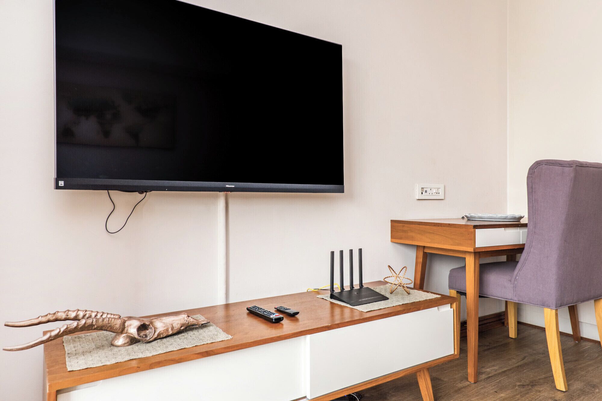 A modern living room with a flat-screen TV mounted on the wall, a wooden coffee table with a remote control, a small sculpture, and a purple chair.