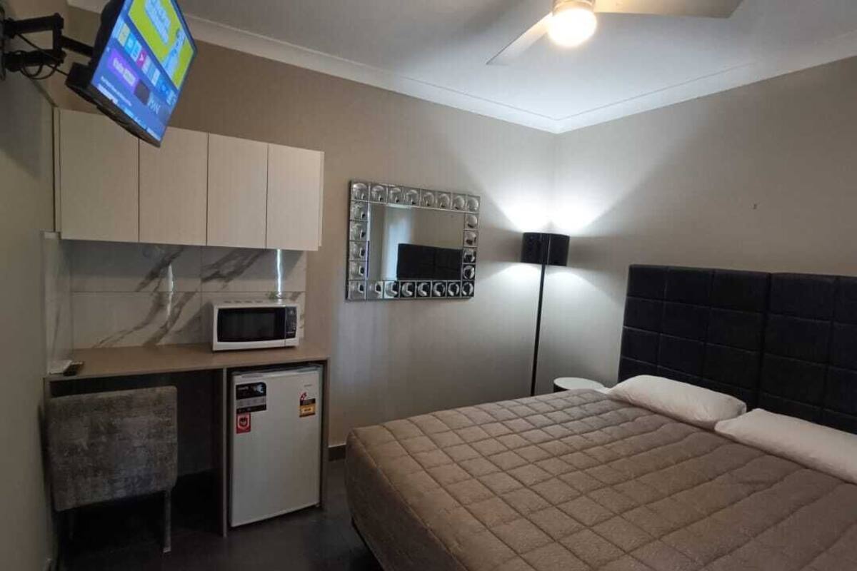 Un dormitorio con cama, escritorio con microondas, refrigerador pequeño, espejo y ventilador de techo.
