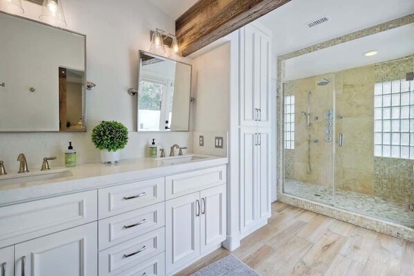 Un baño moderno con un espejo grande, muebles blancos, una planta en maceta y zona de ducha con mampara de cristal.