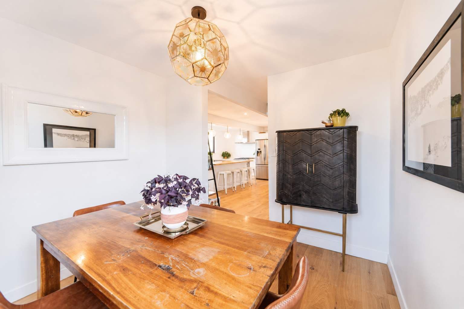 A dining area with a wooden table, chairs, a vase of flowers, a pendant light, and a black cabinet with a chevron pattern.