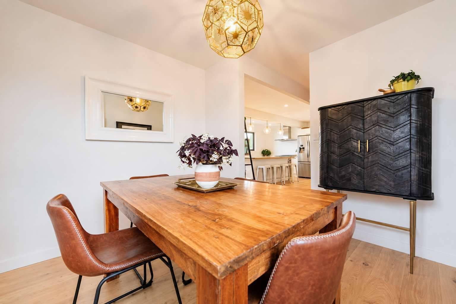 Un coin repas avec une table en bois, des chaises en cuir, un vase de fleurs et un buffet noir avec des poignées dorées.