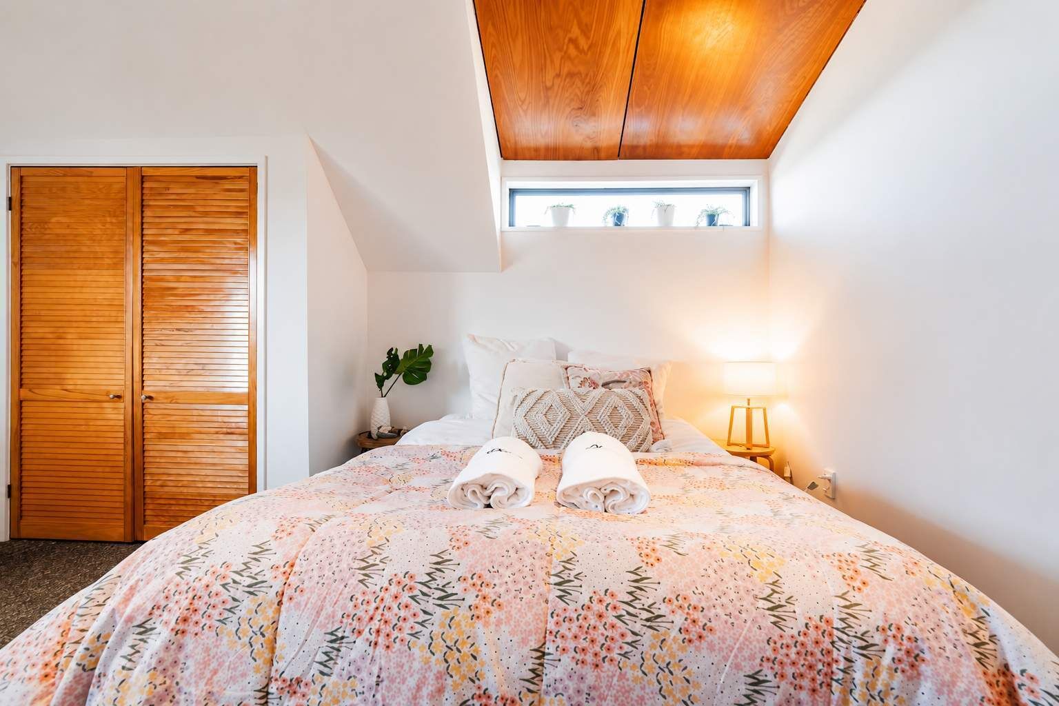 A bedroom with a large bed, a wooden wardrobe, a small bedside table with a lamp, and a window with a view of the sky.