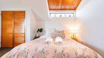 A bedroom with a large bed, a wooden wardrobe, a small bedside table with a lamp, and a window with a view of the sky.