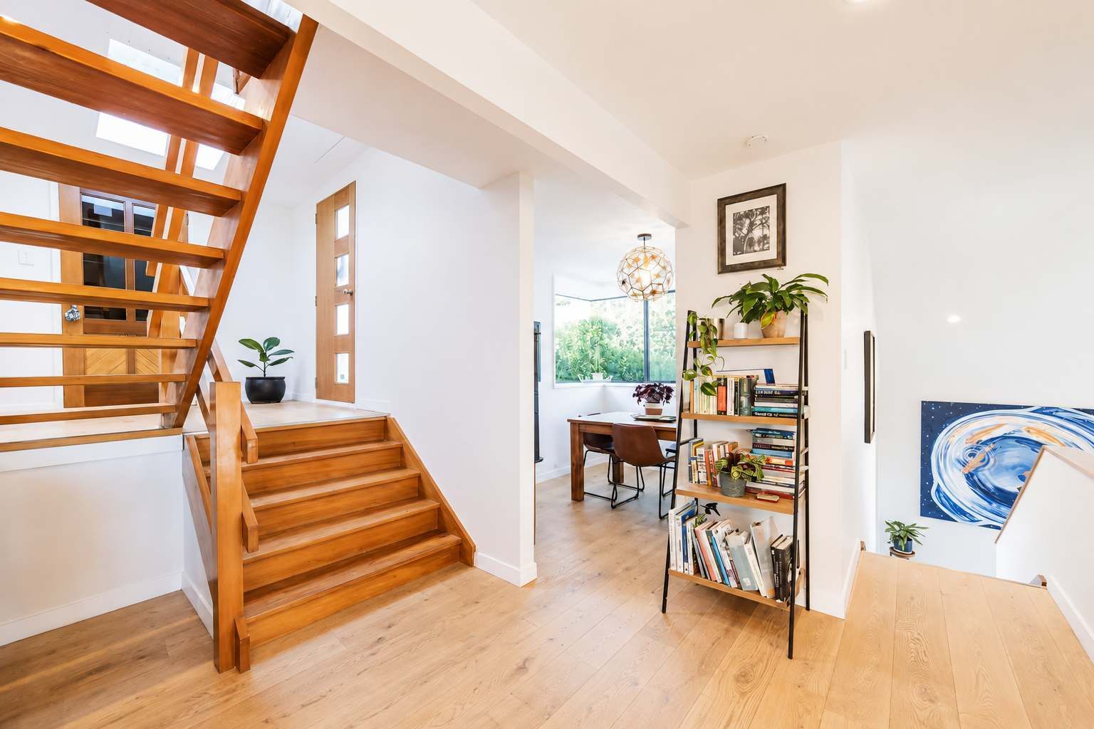Ein modernes Interieur mit einer Holztreppe, einem Schreibtisch mit Stuhl, einem Bücherregal und einem großen Fenster.