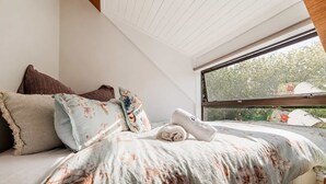 A bedroom with a bed, pillows, a rolled towel, and a window with a view of greenery.