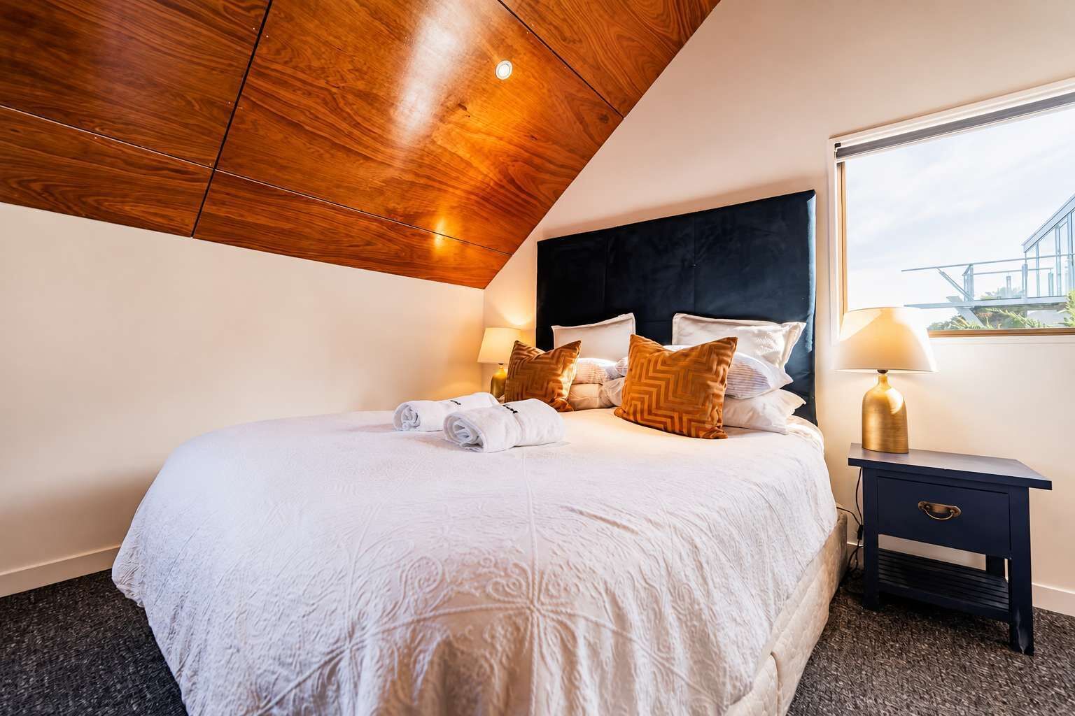 A hotel room with a large bed, a dark headboard, a nightstand with a lamp, and a view of the outside through a window.
