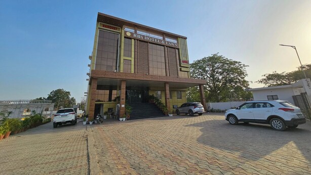A hotel with a prominent 'RBS Hotel & Lawn' sign, a covered entrance, and a paved driveway with parked cars. - RBS Hotel And Lawn (Faizabad)