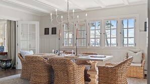 A spacious room with a large dining table surrounded by wicker chairs, a chandelier, and large windows with a view of trees. - Lakeview Bliss - Beautifully Decorated Home! Close Private Beach! (Harbor Springs)
