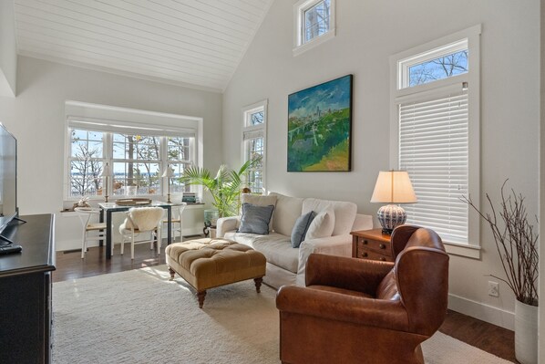 A spacious living room with a high ceiling, a large window with blinds, a painting on the wall, and a cozy seating area with a sofa and ottoman.