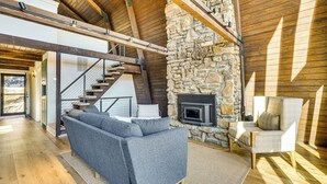 A spacious living area with a stone fireplace, a sofa, and a chair. A staircase with wooden beams leads to an upper level. - Fish, Swim & Kayak: Spacious Beaver Lake Home! (Garfield)