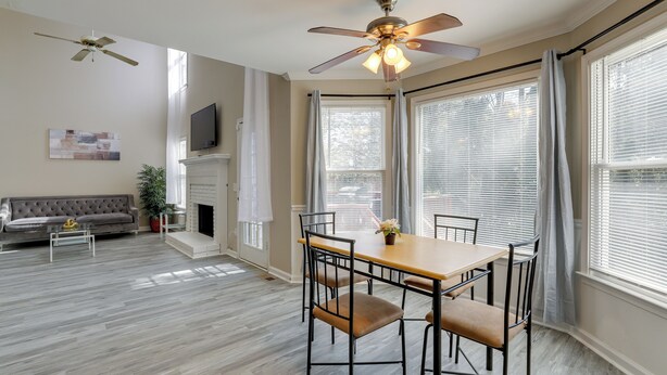 Un espace repas comprenant une table et des chaises, un ventilateur de plafond, une cheminée et de grandes fenêtres équipées de stores.
