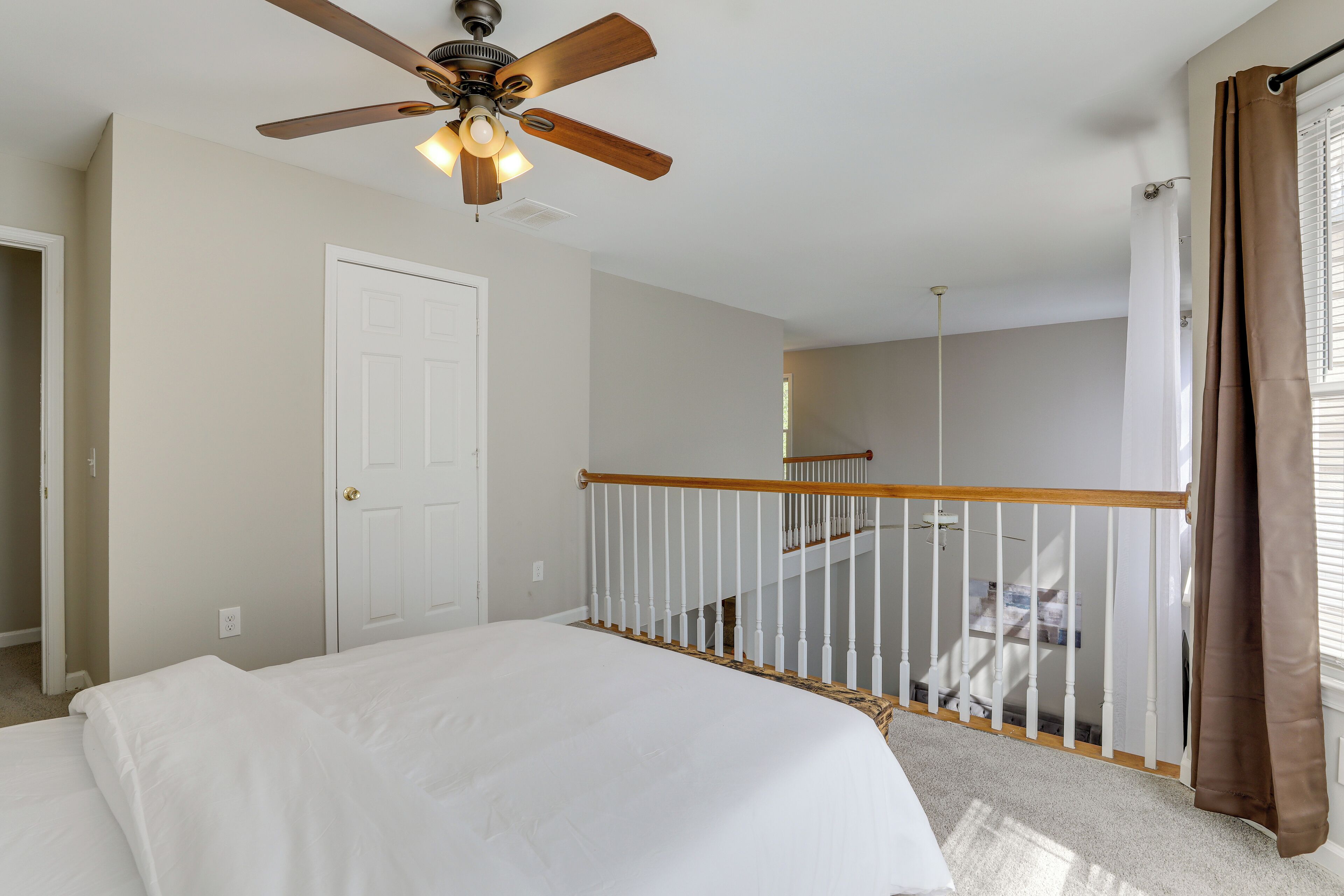 A bedroom with a bed, a ceiling fan, a white door, a staircase with a wooden railing, and a window with curtains.