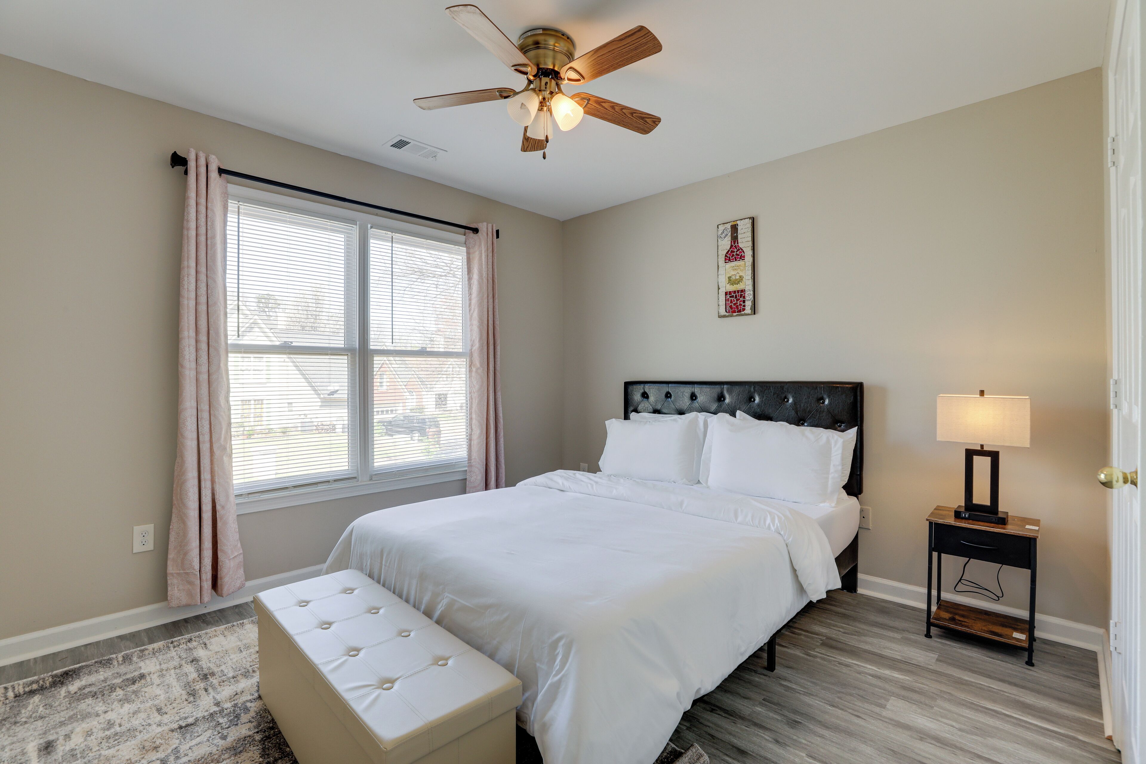A bedroom with a bed, a ceiling fan, a nightstand with a lamp, a window with curtains, and a wall-mounted artwork.