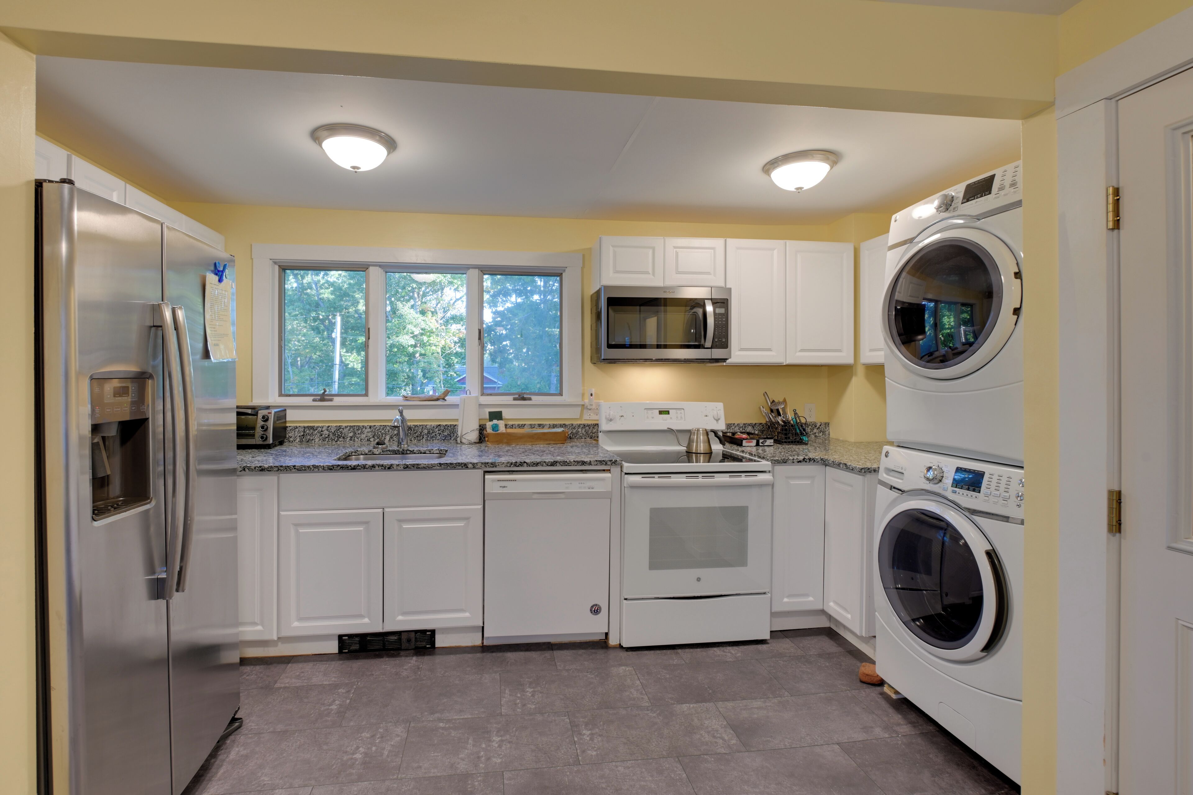 A modern kitchen with white cabinets, a stainless steel refrigerator, a built-in oven, a microwave, and a front-loading washer.