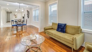 Een ruime woonkamer met houten vloeren, een beige bank, een glazen salontafel en een eethoek met een metalen tafel en stoelen.
