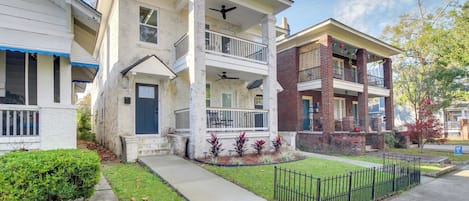 A two-story apartment building with a front porch, a balcony on the second floor, and a small garden area.