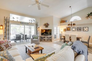A spacious living room with a sofa, coffee table, and a television. There is a dining area with chairs and a table. The room has a ceiling fan, a large window, and a decorative sign on the wall. - Fort Myers Vacation Rental w/ Lanai & Private Pool (Fort Myers)