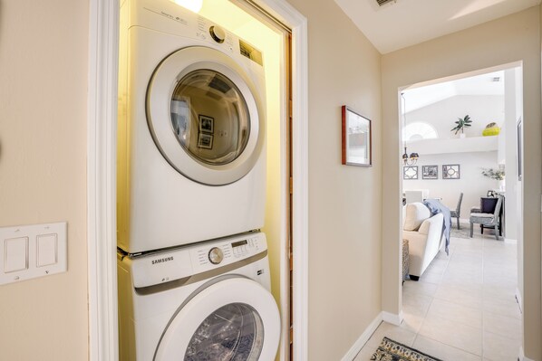 Une buanderie équipée d’un lave-linge et d’un sèche-linge Samsung, et une porte donnant sur un espace de vie comprenant un canapé et une table à manger.