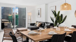 A modern dining area with a wooden table set for four, a black leather sofa, and a view of a cityscape through a large window. - Modern 1BR | Free Parking | Pool | Gym (Brickell)