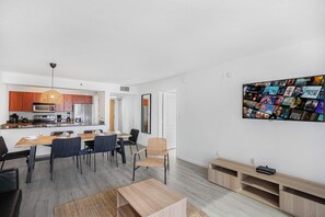 Una cocina moderna con armarios de madera, una zona de comedor con mesa y sillas de madera, y un televisor montado en la pared que muestra carteles de películas.