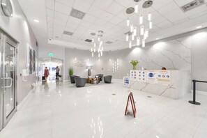 A modern hotel lobby with a clean, polished floor, a reception desk with informational posters, and a seating area with chairs and a small table.