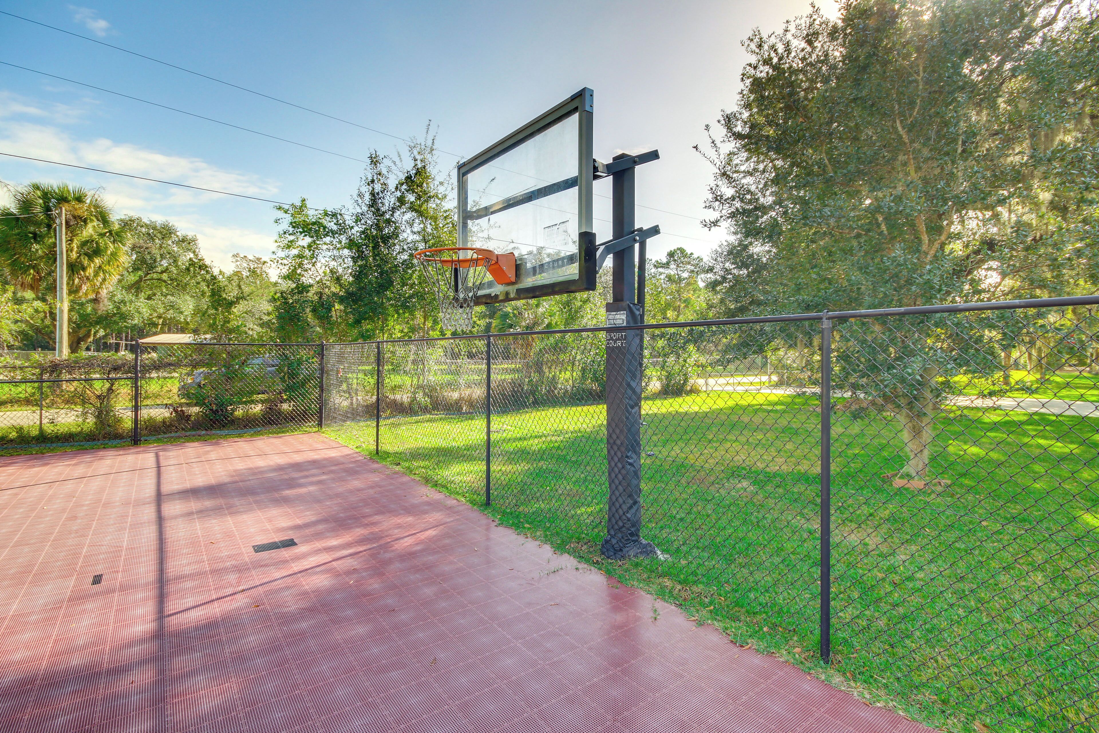 An outdoor basketball court with a metal fence, a basketball hoop, and a red surface.