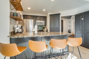 Een moderne keuken met een granieten aanrecht, roestvrijstalen apparatuur en een ingebouwde magnetron. Drie barkrukken en een houten plank met decoratieve objecten.