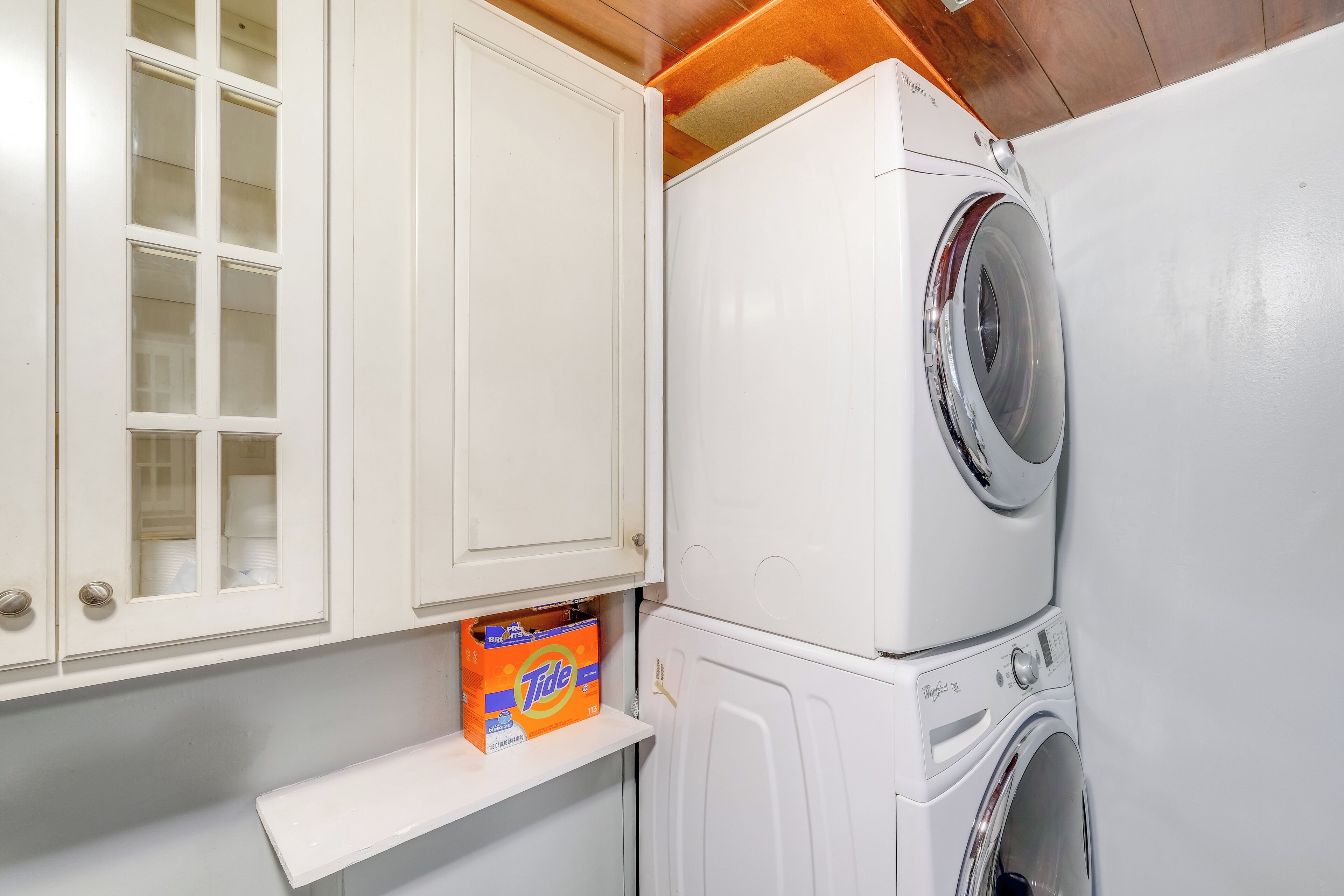 A compact laundry area with a front-loading washer and dryer, a cabinet with a glass door, and a shelf with a Tide detergent box.