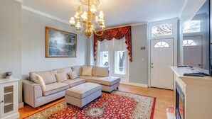 Un salon avec un canapé d’angle, un lustre, un tapis à motifs rouges et un tableau accroché au mur.