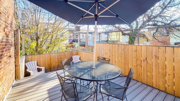 Un patio couvert avec une table ronde et des chaises, une terrasse en bois et une vue sur les arbres et les bâtiments.