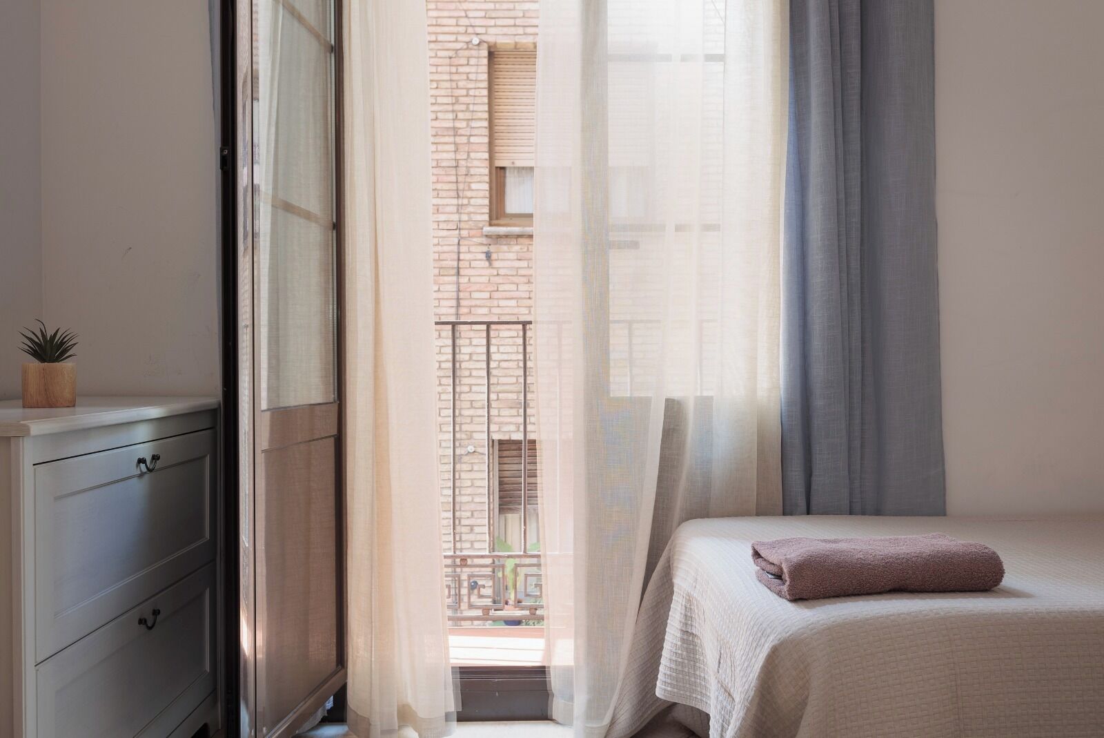 Camera da letto con un letto, una cassettiera, una finestra con tende e vista su un edificio in mattoni.