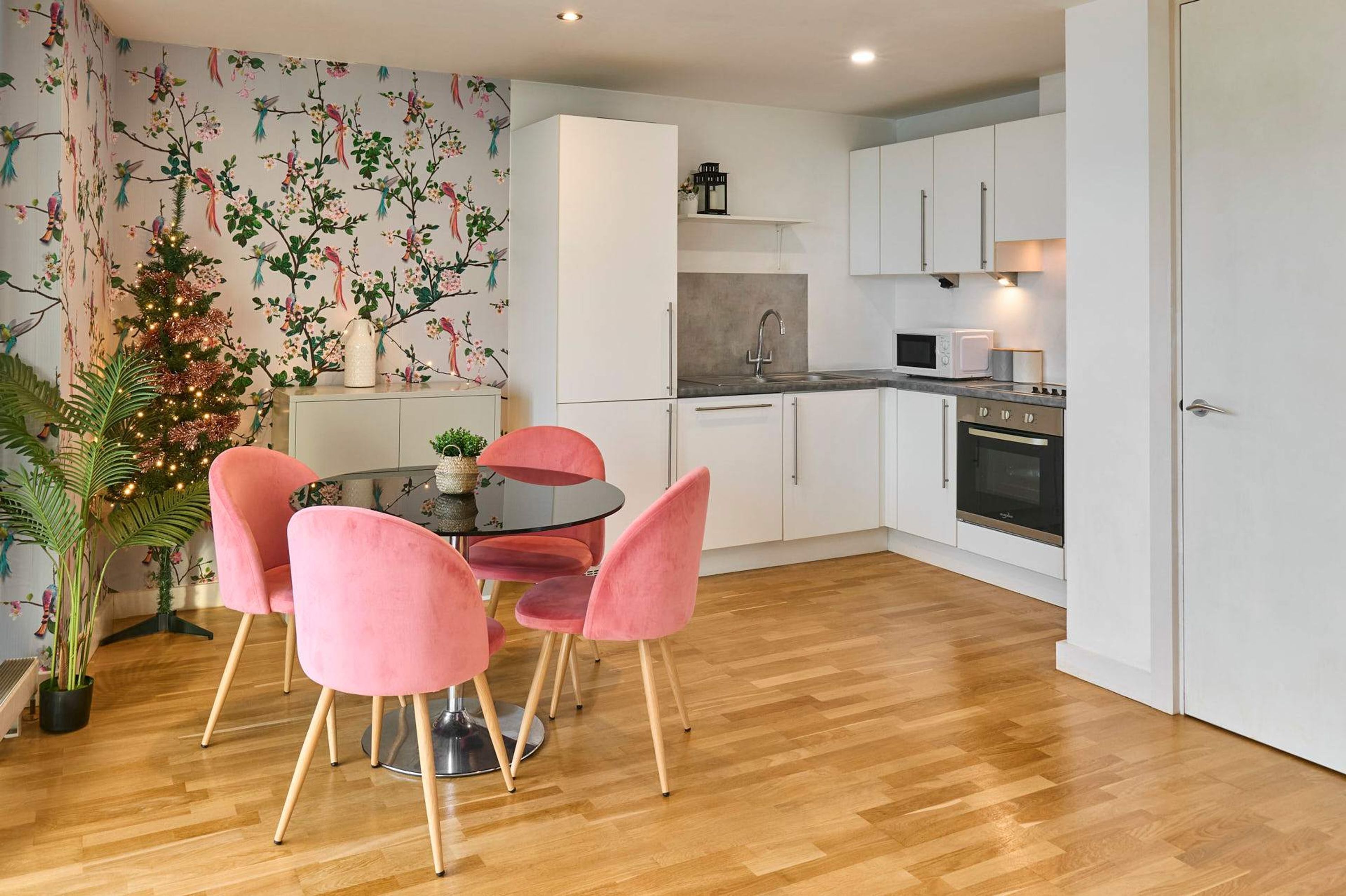 A modern kitchen with white cabinets, a microwave, and an oven. There is a dining area with a round table and pink chairs. A Christmas tree is visible in the corner.