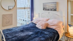 A bedroom with a bed, a round mirror, a framed picture, and a window with a cityscape view.