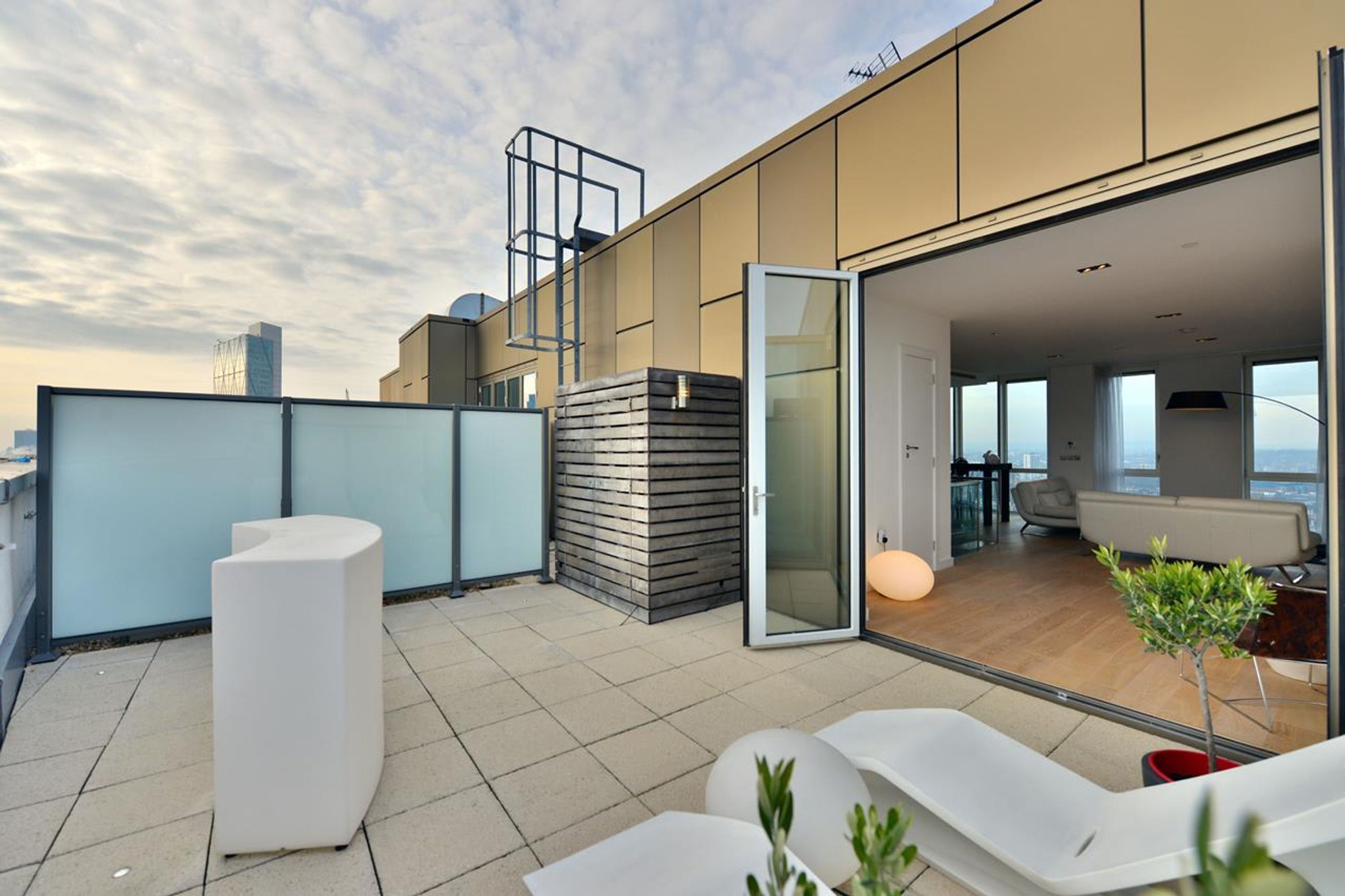 A modern rooftop terrace with a glass door leading to an interior space, furnished with white lounge chairs and a small potted plant.