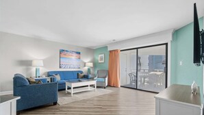 A modern living room with a blue sofa, a white coffee table, and a glass door leading to a balcony. - Ocean View • Beach Across St • Giant Hot Tub/Pool (Myrtle Beach)