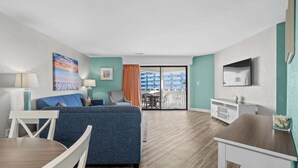A hotel room with a bed, a desk, a chair, a TV, and a balcony with a view of other buildings. - Ocean View   • Beach Across St • Giant Hot Tub/Pool (Myrtle Beach)