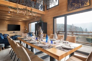 A dining area with a long wooden table set for a meal, surrounded by chairs, with a large window offering a view of the outdoors.