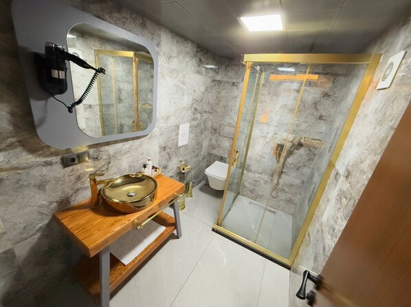 Une salle de bain avec un mur en marbre, un meuble-lavabo en bois avec un évier doré, une douche à parois de verre et un sèche-cheveux fixé au mur.