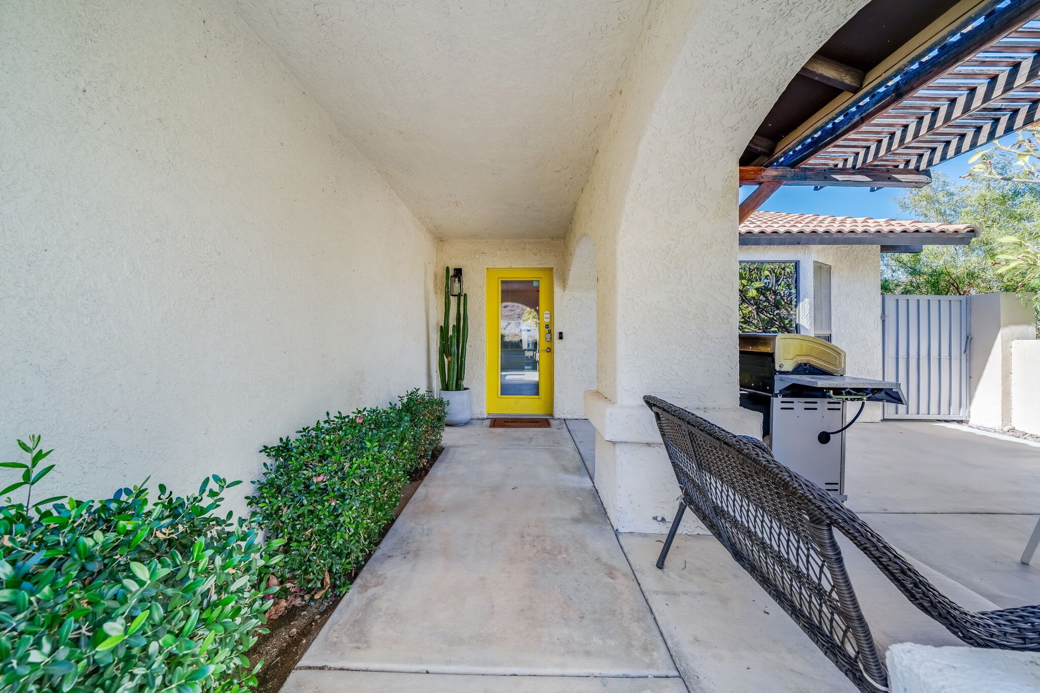 A covered walkway leading to a yellow door, with a grill and outdoor seating area on the right.