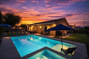A swimming pool with lounge chairs and umbrellas, surrounded by a house with a tiled roof and a well-lit patio area. - Pet Friendly | Private Pool, Spa, Fire Pit, Patio (Indio)