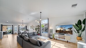 Een moderne woonkamer met een grijze bank, een salontafel en een televisie op een witte kast. Er is een zithoek met een tafel en stoelen, en een trap die naar een verdieping leidt.