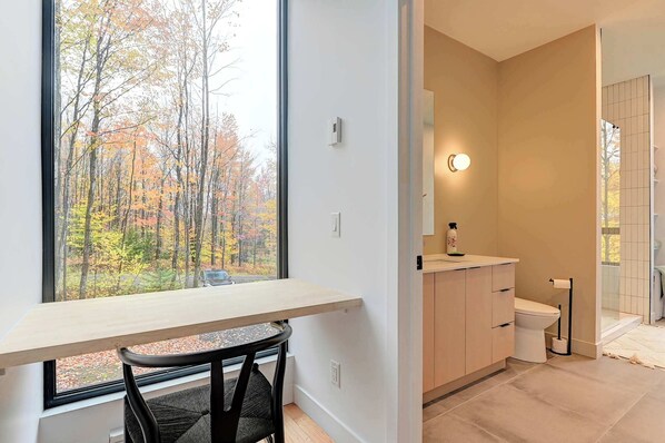 A modern bathroom with a toilet, sink, and glass shower. A window offers a view of autumn trees. - Chalet Black Box (Mansonville)