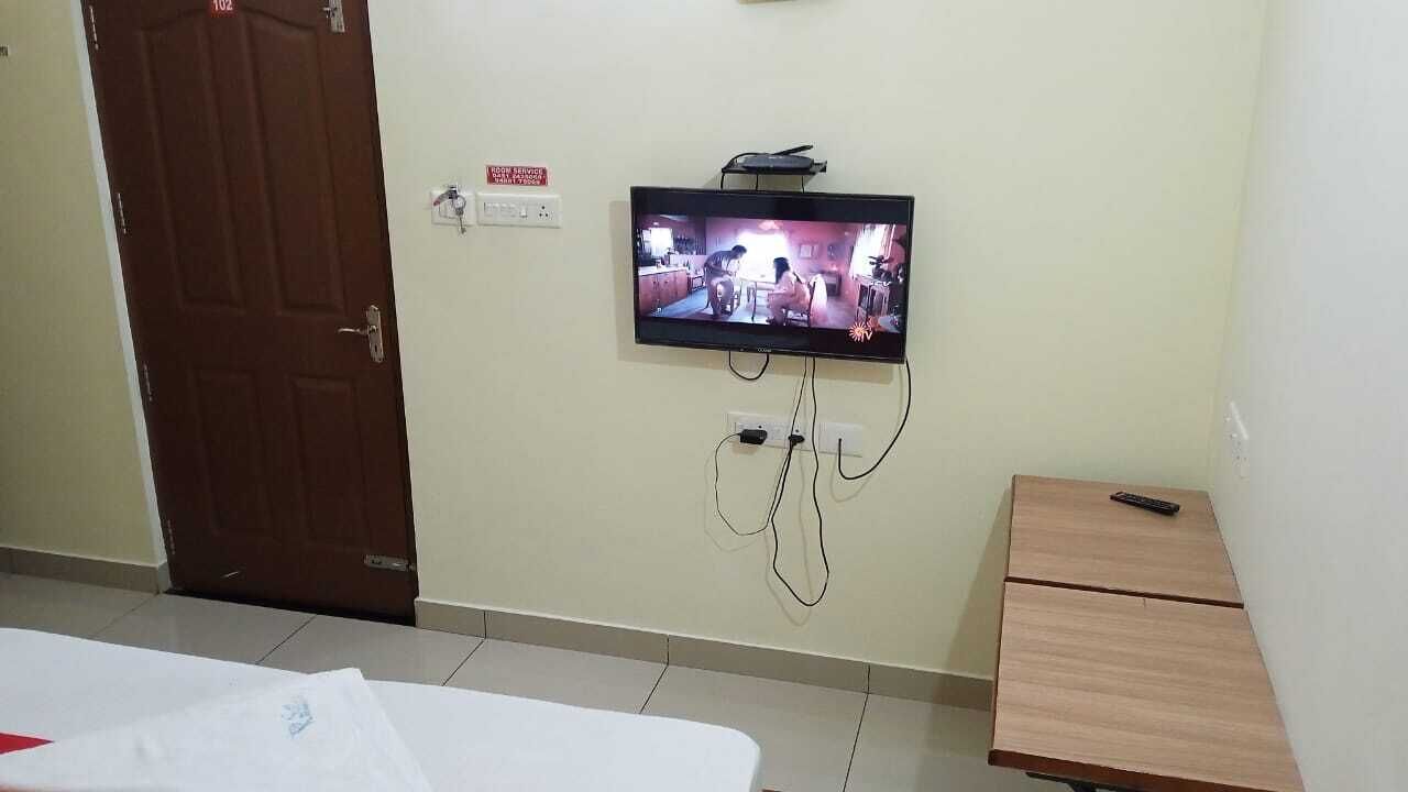 A room with a television mounted on the wall, a wooden cabinet, and a bed with a white sheet.