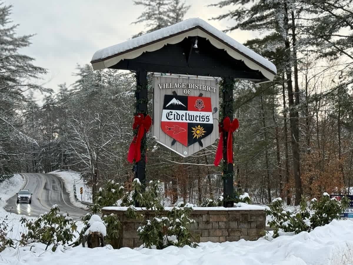 A snow-covered village sign with a mountain and sun emblem, surrounded by trees.