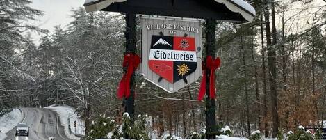 A snow-covered village sign with a mountain and sun emblem, surrounded by trees.