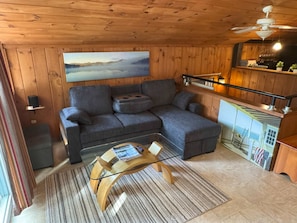 A cozy cabin interior with a sectional sofa, a wooden ceiling, a painting of a landscape, a small table with a book, and a kitchenette area. - Serene Forest Chalet - Beaches | Kayaks | Bikes (Madison)