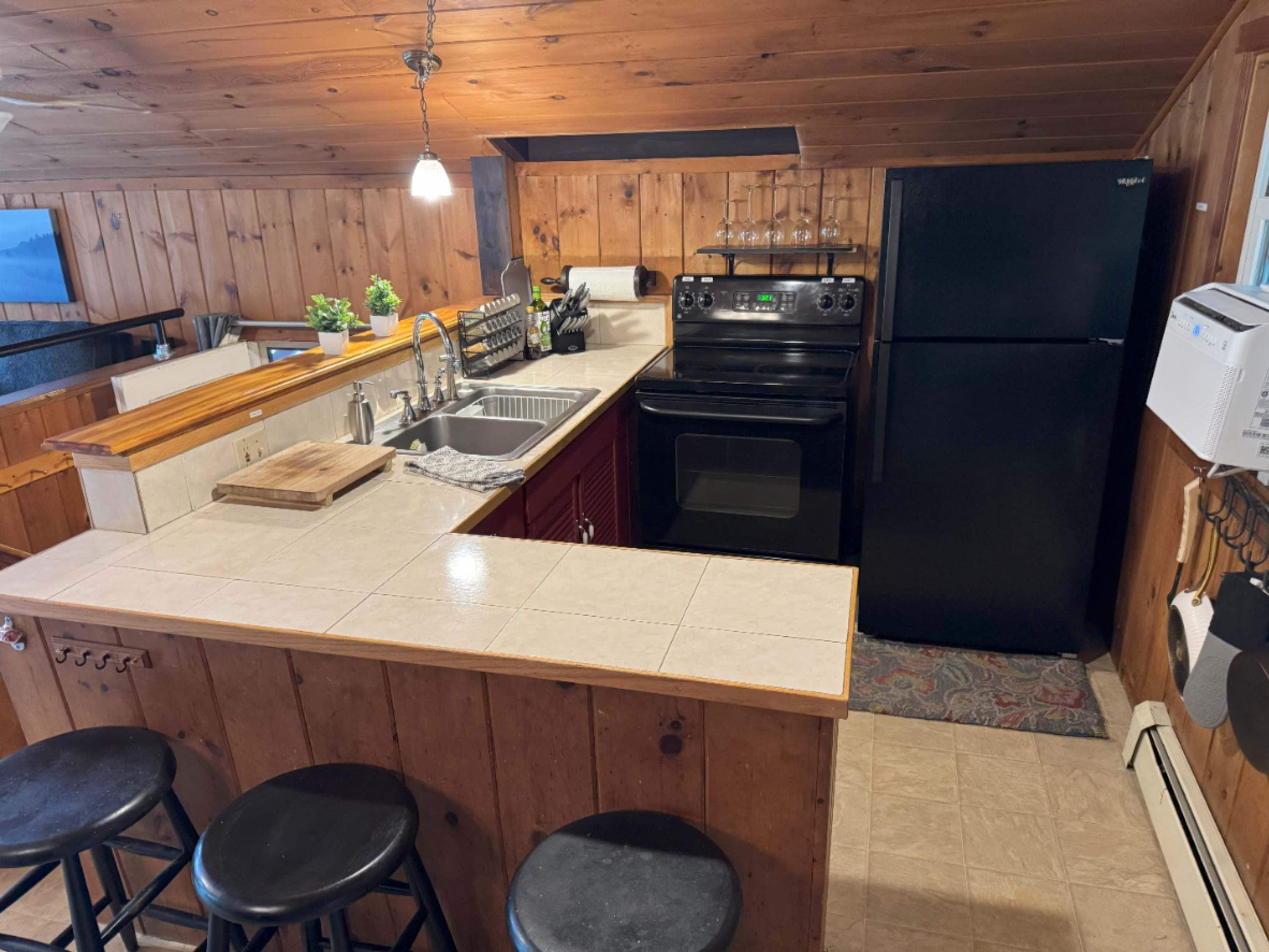 Una cocina con paredes de madera, un refrigerador negro, una estufa, un fregadero y taburetes de bar.