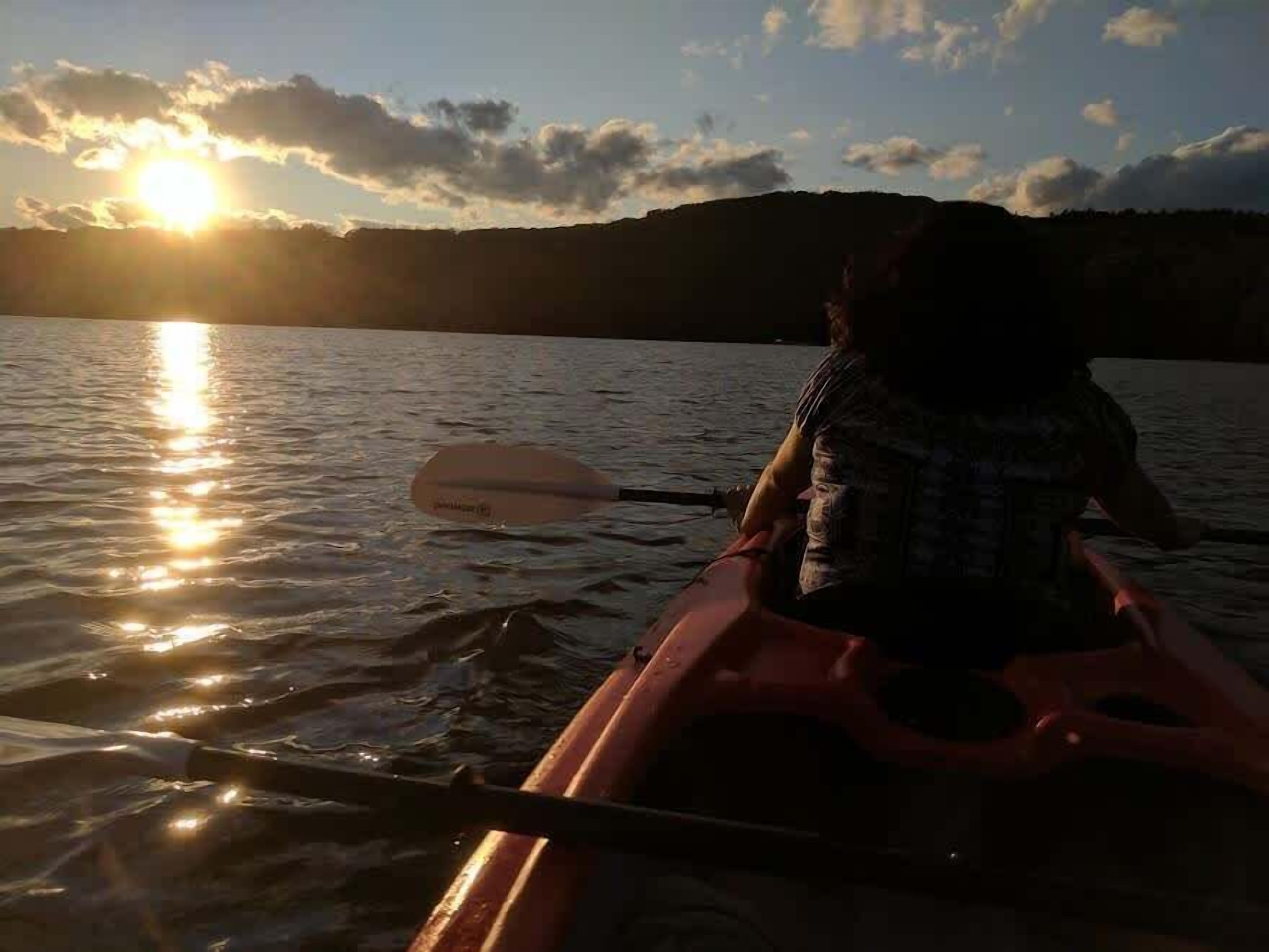 A person in a kayak on a body of water with the sun setting in the background.
